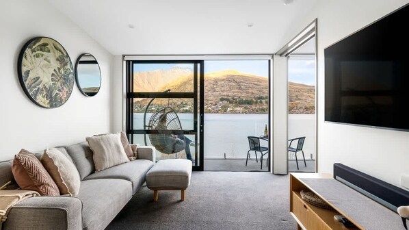 A modern living room with a sofa, a chair, a flat-screen TV, and a view of a lake and mountains.