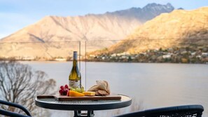 Une table avec une bouteille, du fromage, des raisins et du pain, donnant sur un lac avec des montagnes en arrière-plan.
