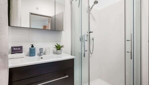 Une salle de bain moderne avec une douche à l’italienne en verre, un lavabo blanc avec un miroir au-dessus et un meuble de rangement de couleur foncée en dessous.