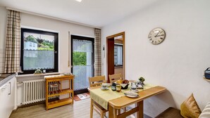 Un coin repas avec une table dressée pour deux personnes, une horloge au mur et une vue sur une maison par la fenêtre.