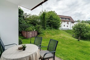 A balcony with a table and chairs, overlooking a grassy area and a white building.