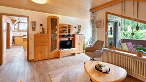 Una sala de estar moderna con un mueble bajo de televisión de madera, un sillón cómodo y un ventanal con vistas a zonas verdes.