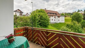 Un balcon donnant sur un quartier résidentiel, une table recouverte d’une nappe verte, un pot de fleurs et une balustrade en bois.