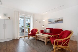Un salon avec un canapé rouge, deux fauteuils en osier, une table basse en bois, une lampe et un tableau accroché au mur.