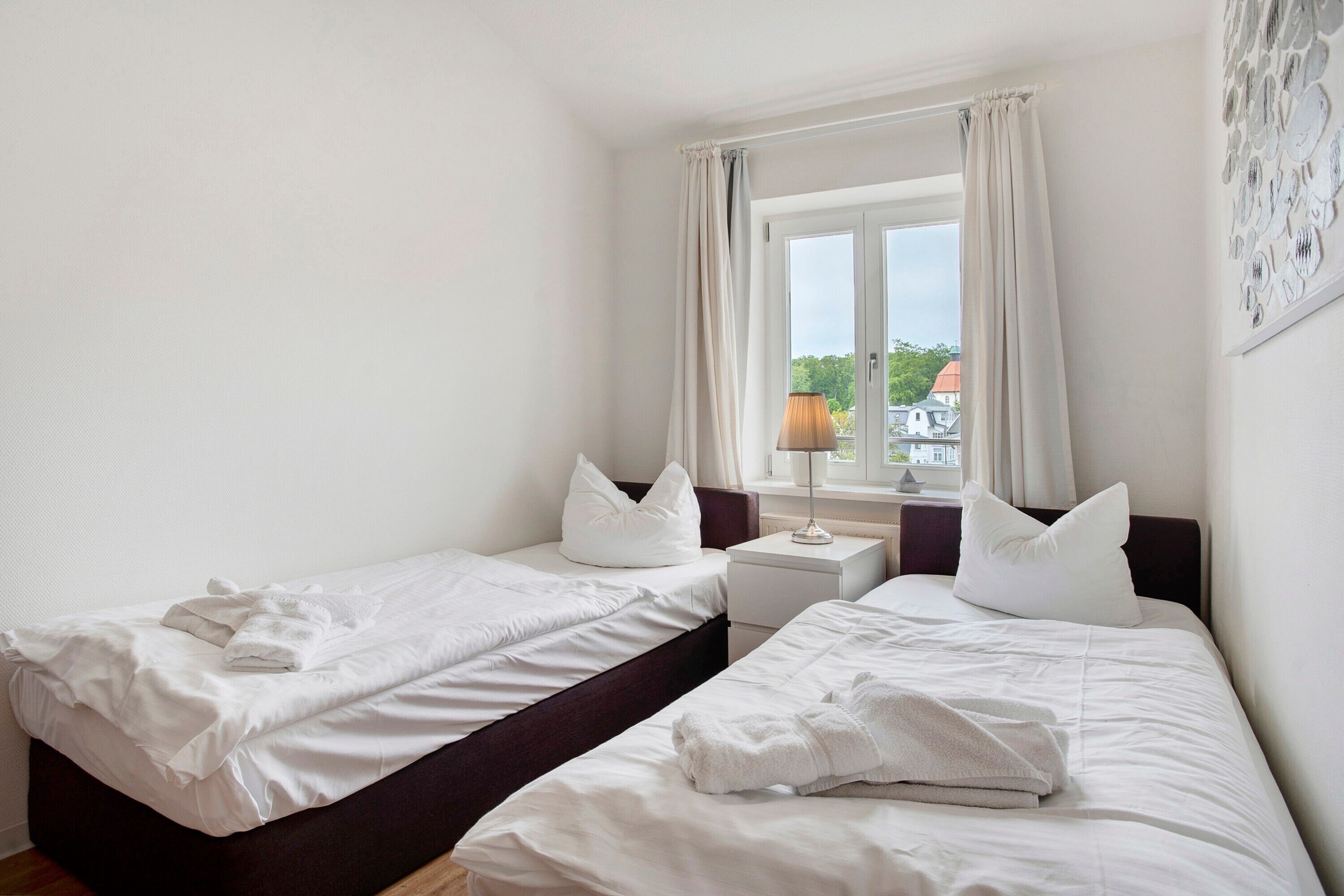 A room with two single beds, a window with curtains, a nightstand with a lamp, and a wall with a plate pattern.