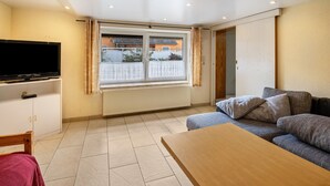 A living room with a sofa, a TV, a wooden table, and a window with a view of buildings. - Apartment 'Ruhiges Nest Mit Sauna Und Garten' with Shared Garden and Wi-Fi (Jessen (Elster))