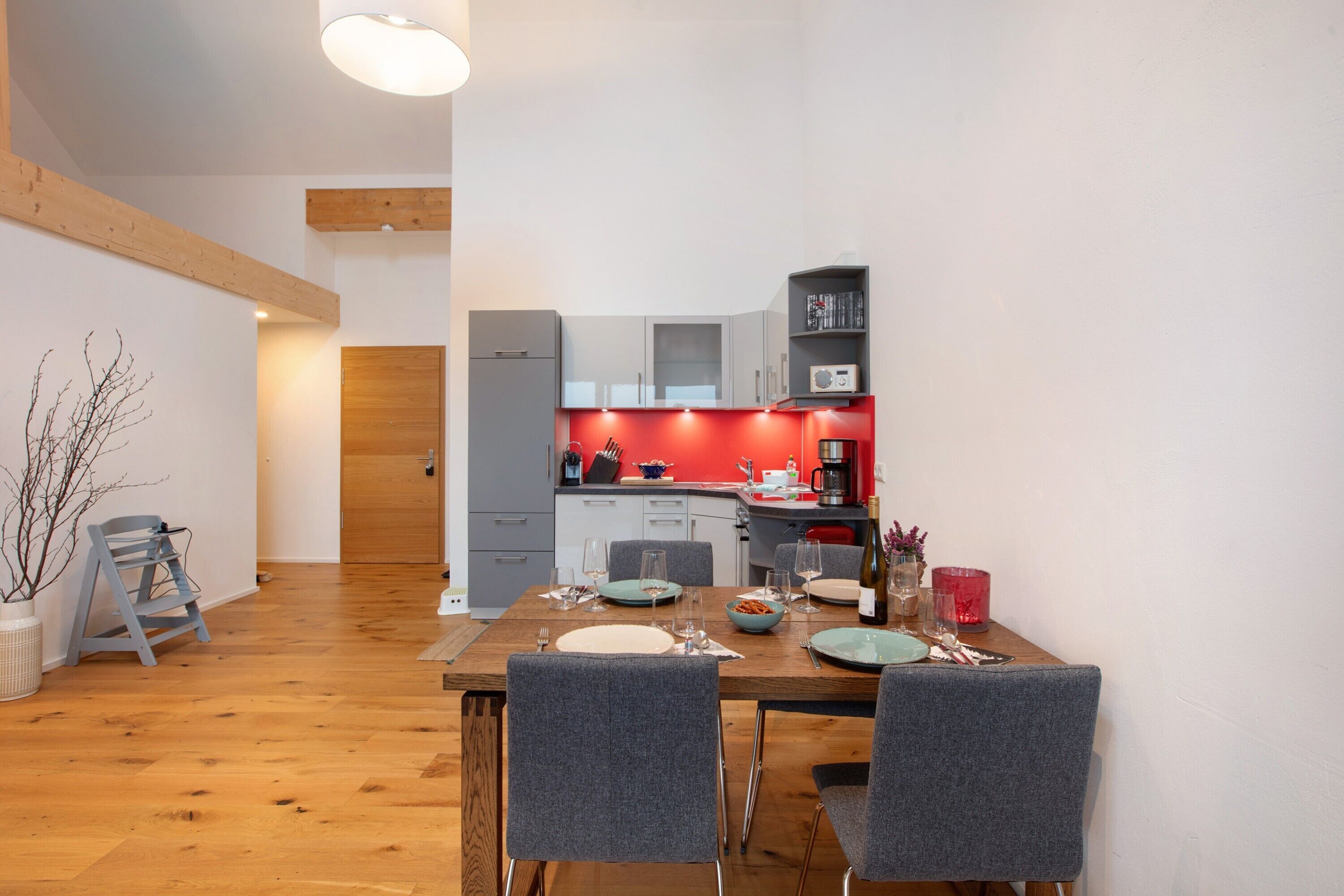 Eine moderne Küche mit grauen Schränken, einer roten Akzentwand und einem Essbereich mit grauen Stühlen und einem Holztisch.