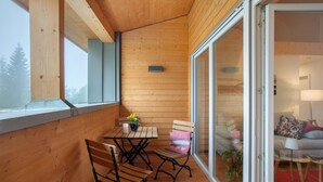 A wooden balcony with a table and chairs, a small plant, and a sliding glass door leading to an interior space with a sofa and lamp. - Apartment 'Hedi' with Mountain View, Balcony and Wi-Fi (Feldberg (Schwarzwald))