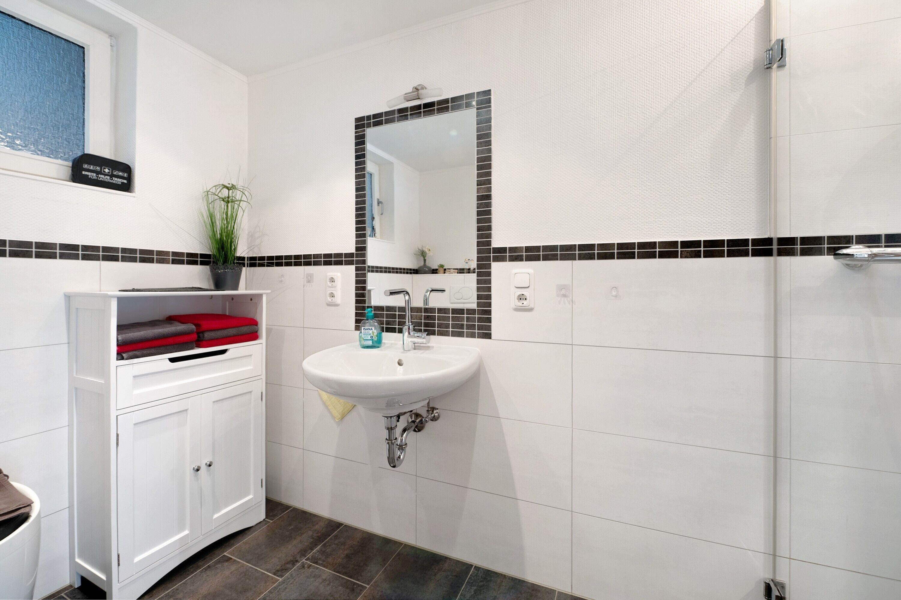 Une salle de bain avec un lavabo blanc, un miroir, une plante en pot et un meuble contenant une serviette rouge.