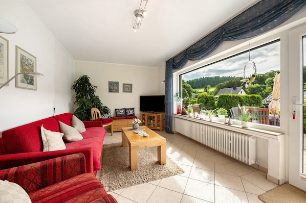 Una sala de estar con un sofá rojo, una mesa de centro de madera y un televisor. Hay una ventana grande con vista a follaje y una casa.