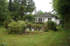 A white house with a tiled roof, surrounded by lush greenery and a pond.