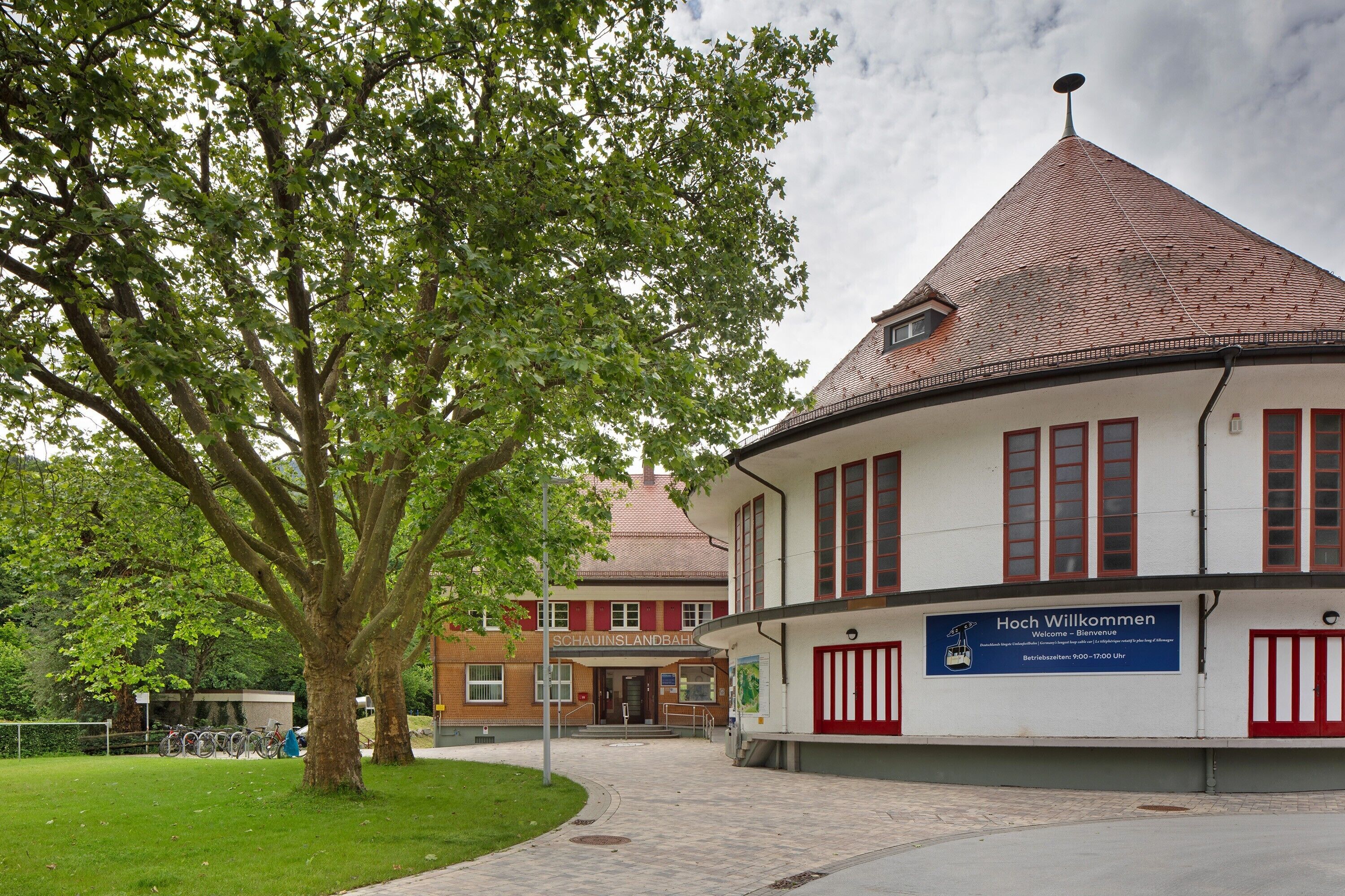 Een gebouw met een rood pannendak, een bord waarop staat 'Hoch Willkommen' en een boom op de voorgrond.
