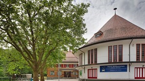 Ein Gebäude mit rotem Ziegeldach, einem Schild mit der Aufschrift 'Hoch Willkommen' und einem Baum im Vordergrund.