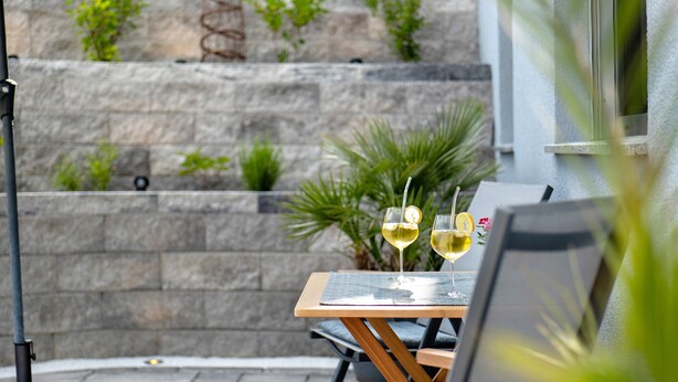 Eine Terrasse mit Tisch, zwei Stühlen und zwei Gläsern Weißwein, vor einer Steinwand mit Pflanzen.