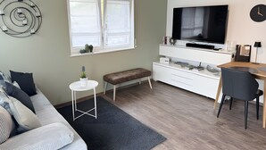 Ein modernes Wohnzimmer mit einer Couch, einem kleinen Tisch mit Pflanze, einer Bank, einem Fernseher und einem Schreibtisch mit Stuhl.