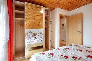 Una camera da letto con soffitto in legno, un letto con copripiumino floreale, un armadio pieno di vestiti appesi e una porta che dà su un'altra stanza.