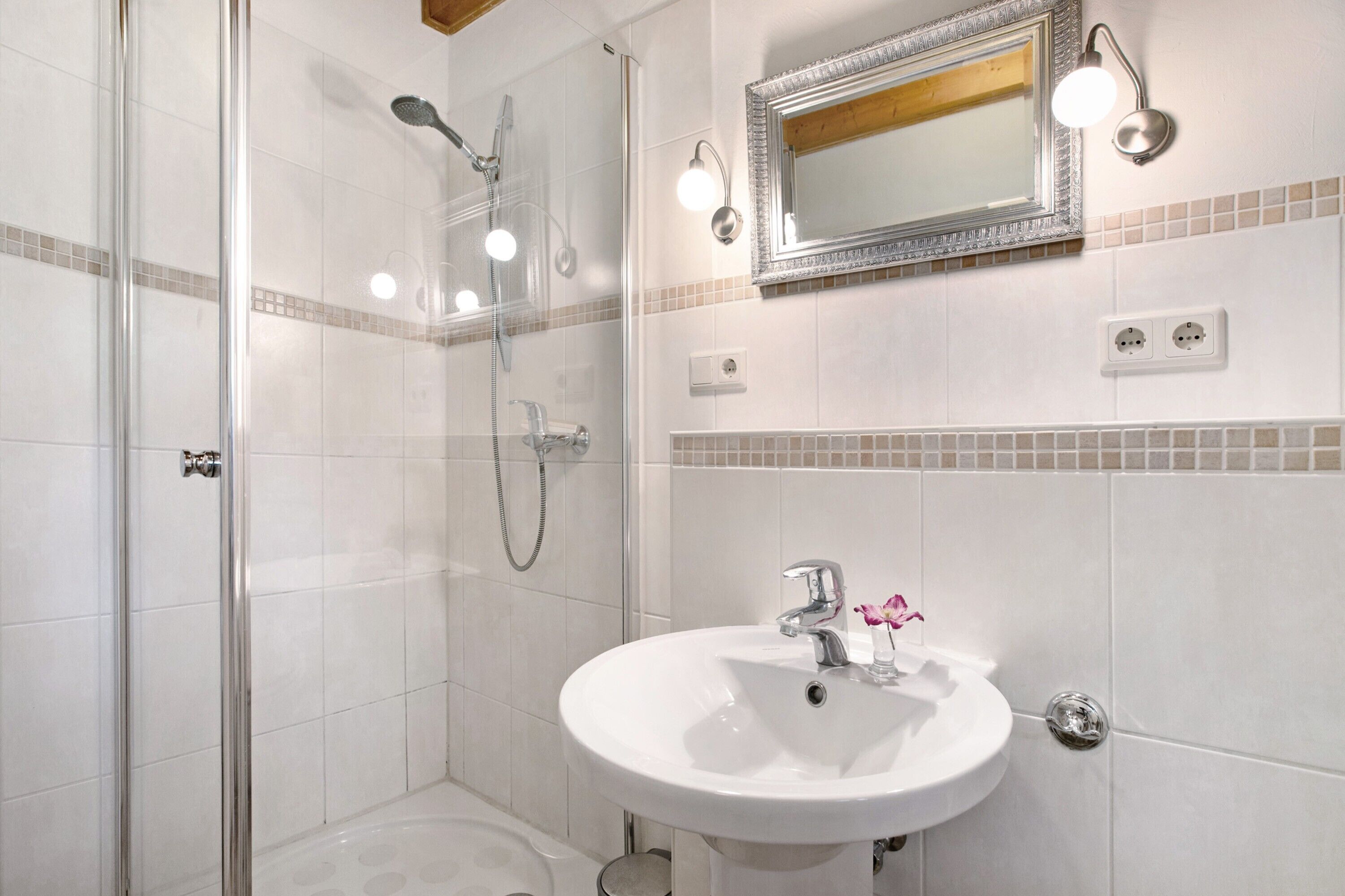 Un bany amb dutxa tancada amb vidre, un lavabo blanc amb un mirall a sobre i una paret amb rajola i una decoració ornamental.