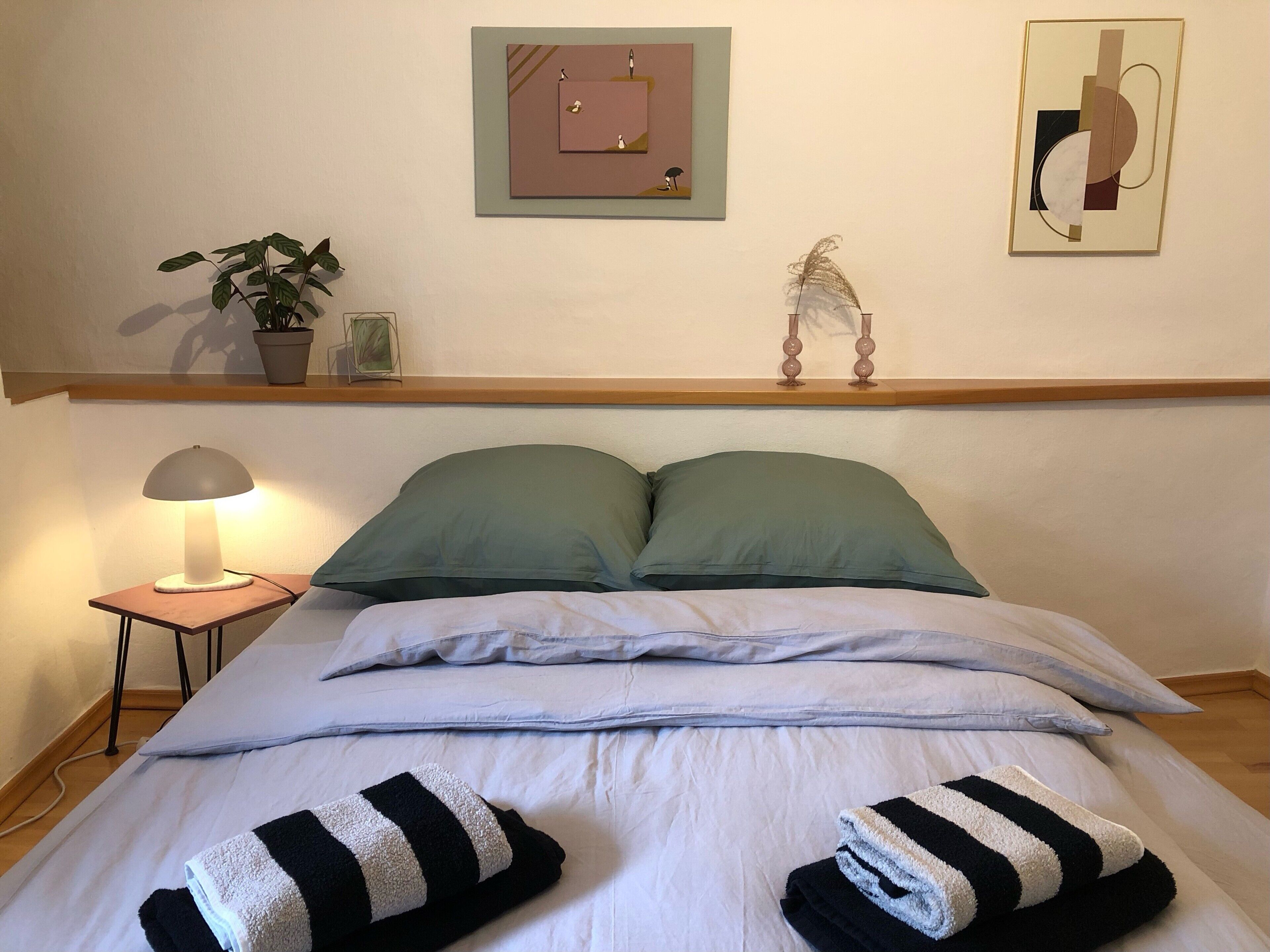 Een net opgemaakt bed met kussens en handdoeken, een bedlamp, een plant en twee decoratieve wandobjecten.