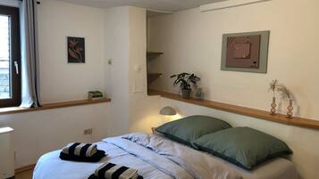 A bedroom with a bed, pillows, a bedside table, a window with curtains, a wall-mounted shelf with a plant, and a framed picture.