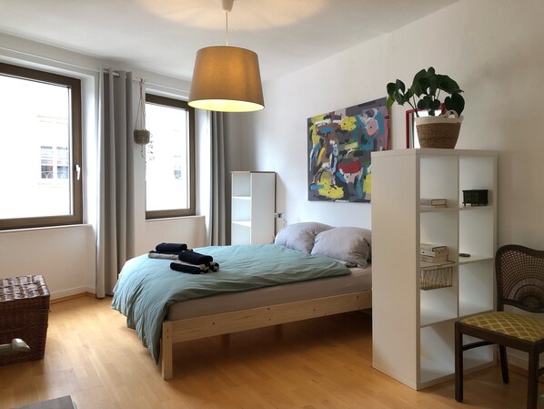 A bedroom with a bed, a hanging light, a window, a plant, a bookshelf, and a chair.