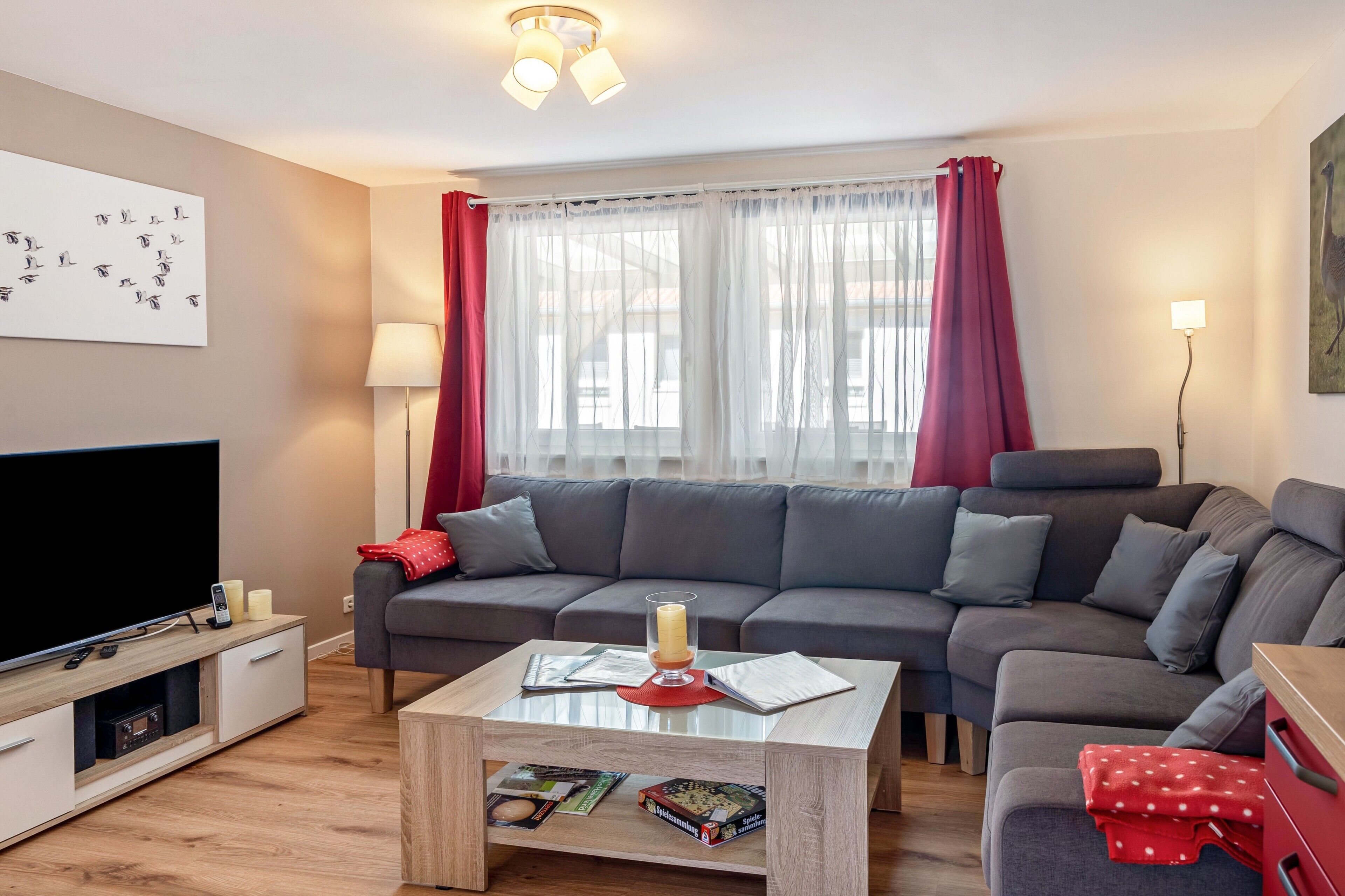 A modern living room with a flat-screen TV, grey sofa, coffee table, and a large window with sheer curtains.