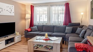 Ein modernes Wohnzimmer mit Flachbildfernseher, grauem Sofa, Couchtisch und einem großen Fenster mit durchsichtigen Vorhängen.