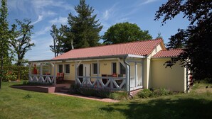 Ein eingeschossiges Haus mit rotem Ziegeldach, einer Veranda mit weißen Geländern und einem kleinen Garten.