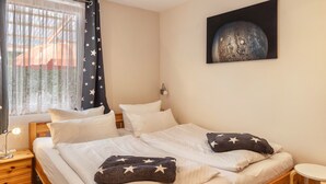 Un dormitorio con dos camas, almohadones estampados de estrellas, una mesita de noche con lámpara, una ventana con cortinas decoradas y un cuadro colgado en la pared.