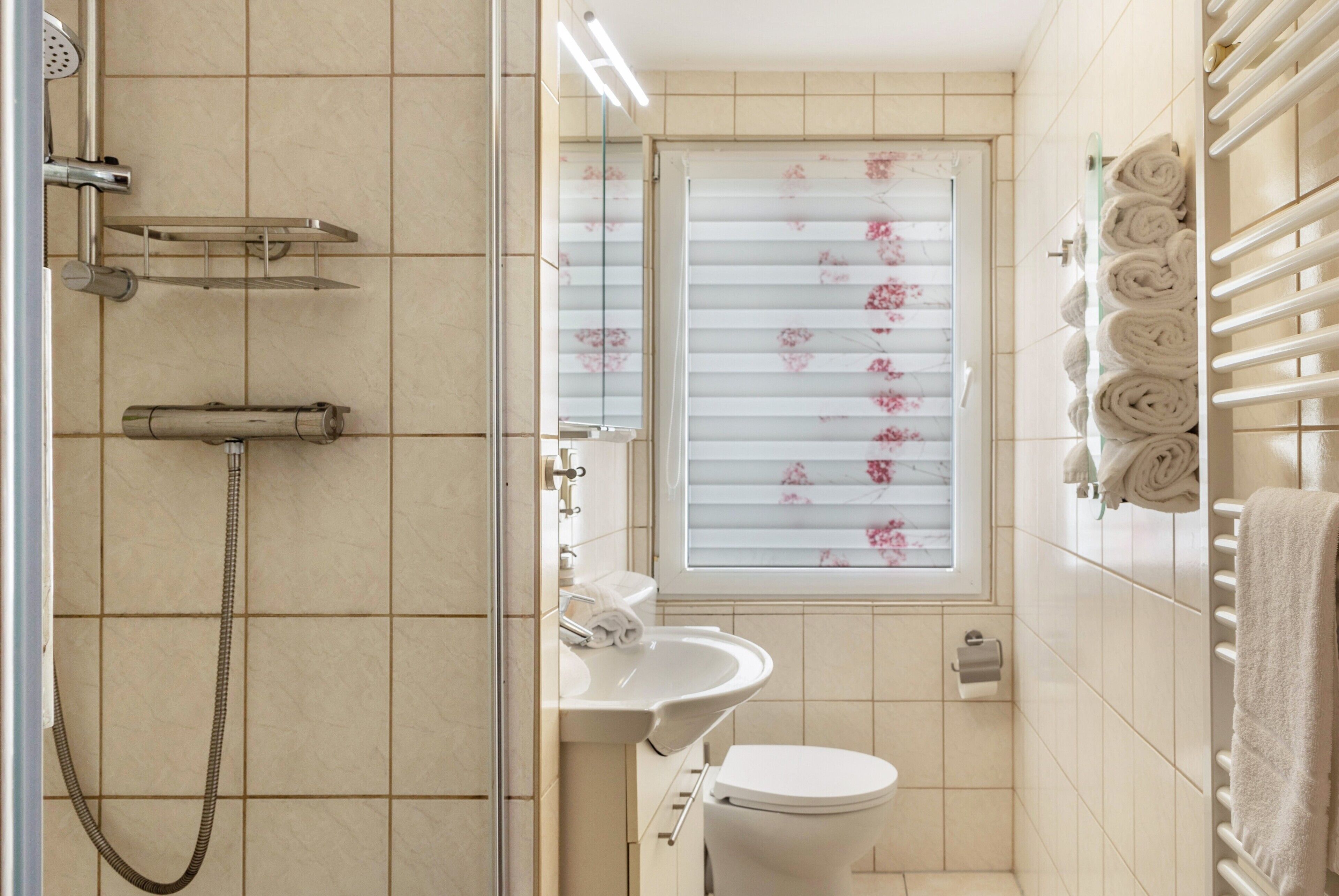 Ein Badezimmer mit Dusche, Toilette, Waschbecken und einem Fenster mit floralem Sichtschutz.