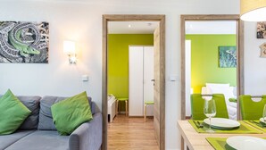 Habitación de hotel moderna con un sofá gris, cojines verdes, una mesa de comedor preparada para dos personas y una pared de acento verde.