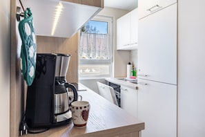 Une cuisine compacte équipée d’une machine à café, d’une tasse, d’une fenêtre avec vue et de meubles blancs.