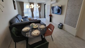 En moderne stue med spiseplads, en grå sofa, et rundt spisebord dækket med tallerkener og bestik, samt et stort tv på væggen.