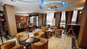 Un hall de hotel con mostrador de recepción, zona de estar con sillones y mesas, diseños decorativos en la pared y ventanas amplias con cortinas.