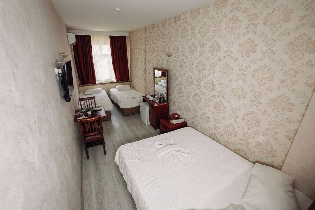Quarto de hotel com cama, escrivaninha, cadeira, mesinha, espelho e janela com cortinas.