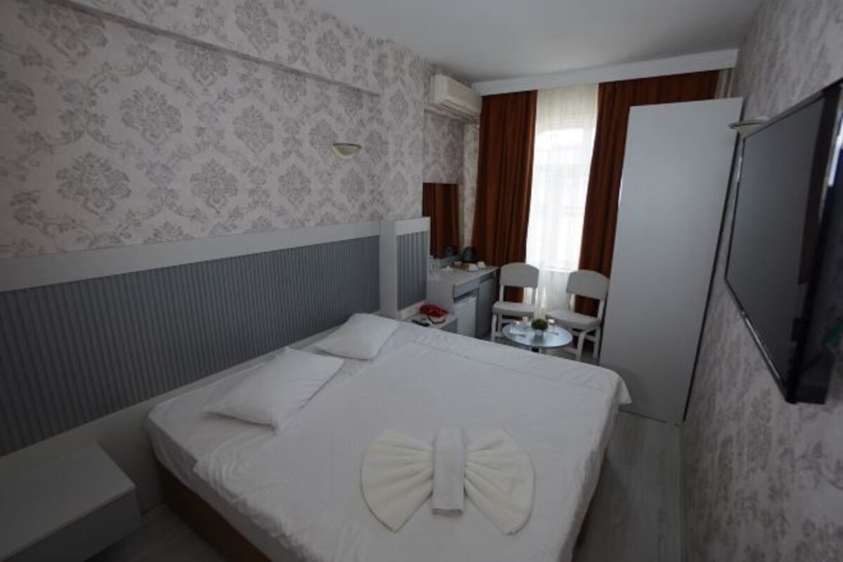 A hotel room with a neatly made bed, a small desk with a chair, a television, and a window with curtains.
