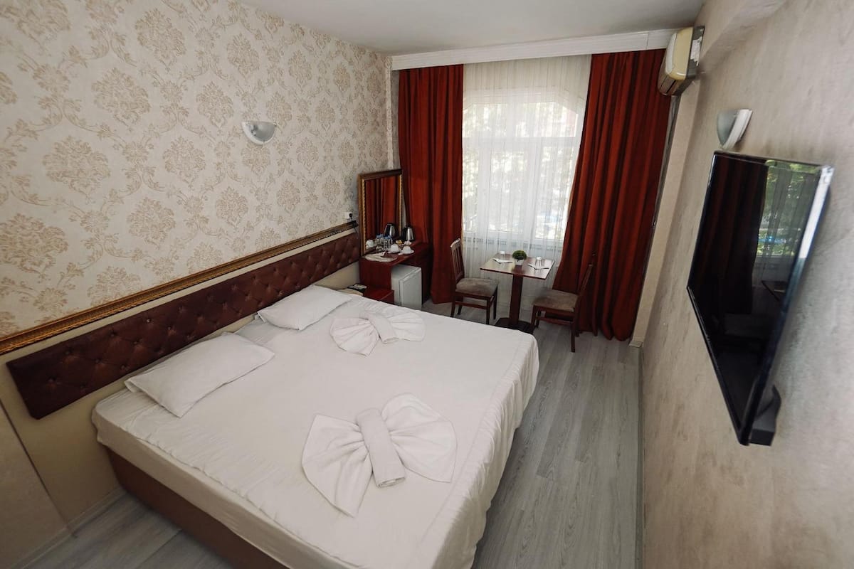 Quarto de hotel com uma cama grande, uma escrivaninha, uma cadeira, uma televisão e uma janela com cortinas.
