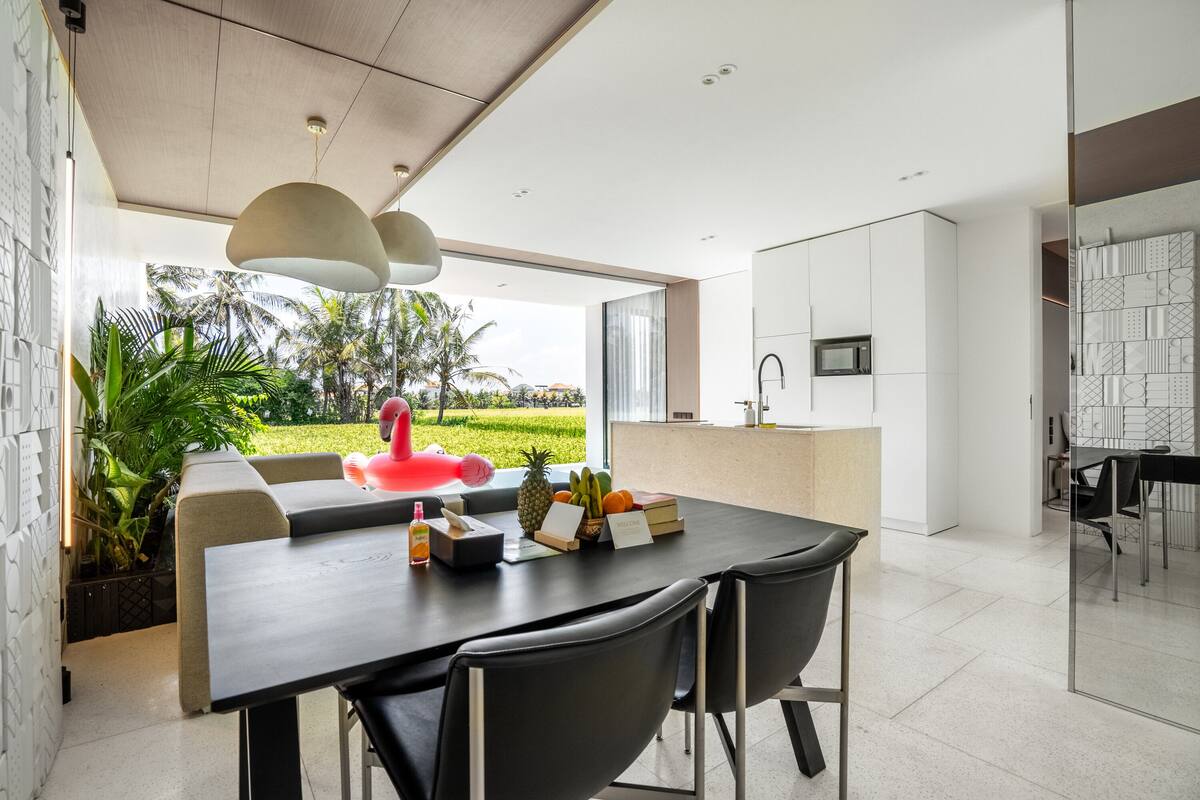 A modern dining area with a large table, chairs, and a view of a garden with a flamingo statue.