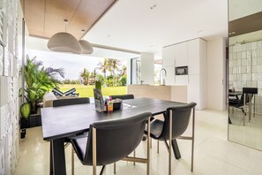 Un comedor moderno con una mesa de madera oscura y sillas negras, una zona de cocina con armarios blancos y vistas a un jardín con palmeras.