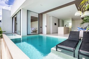 Un área de piscina moderna con agua azul cristalina, sillones de descanso negros y una sala con paredes de vidrio y interior blanco.