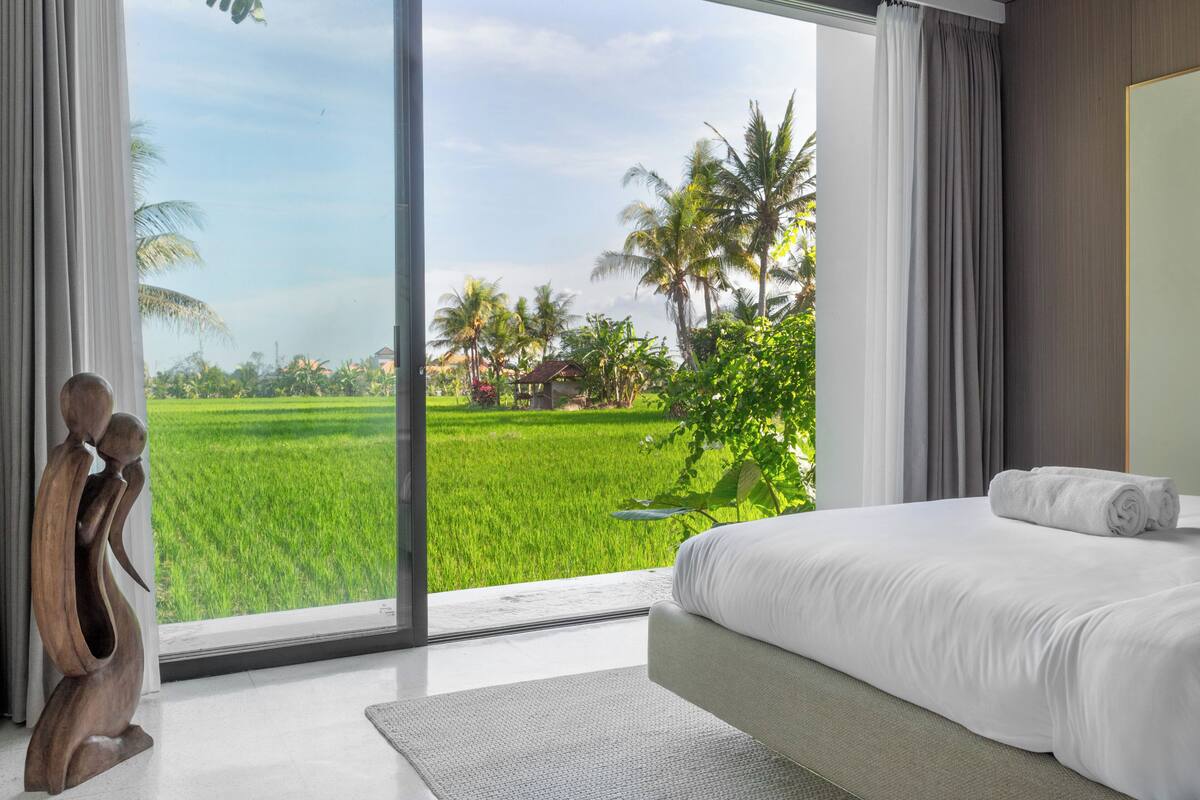 A bedroom with a large window offering a view of a green field and palm trees.