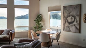 Un salon moderne doté d’une grande fenêtre donnant sur un lac, un coin repas avec une table ronde et un tableau abstrait accroché au mur.