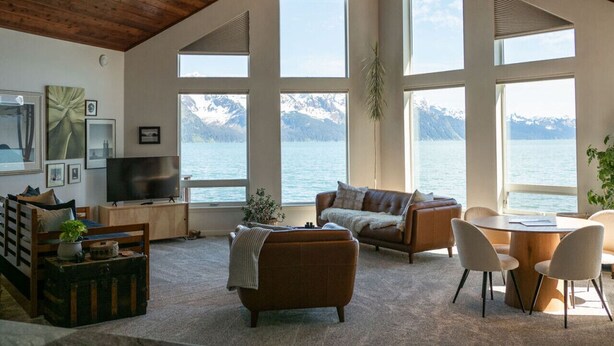Un vaste salon doté d’un haut plafond, de grandes fenêtres offrant une vue sur les montagnes et un lac, un coin salon confortable avec une cheminée, et un téléviseur posé sur un meuble en bois.