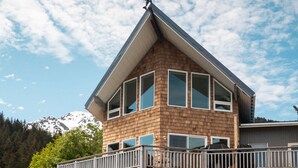 Une maison à deux étages avec un balcon, de gros rochers à l’avant et des montagnes en arrière-plan.