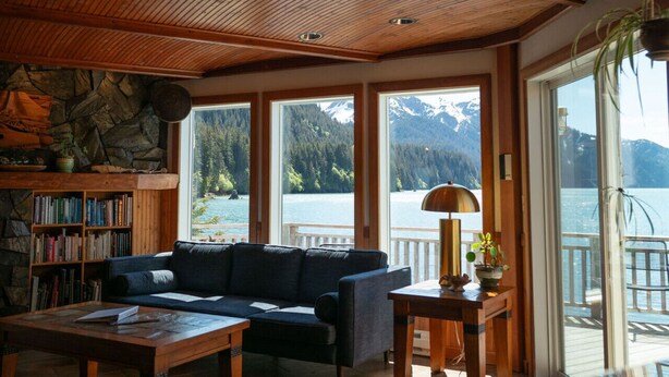 Un salon chaleureux avec un canapé, une table basse, une lampe, une bibliothèque et une vue sur un lac, avec des montagnes en arrière-plan.