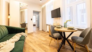 A modern living room with a green sofa, a round wooden dining table with chairs, a flat-screen TV mounted on the wall, and large windows with sheer curtains. - Estrella 8 Wypoczynek i Wygoda Toru Stare Miasto (Torun)