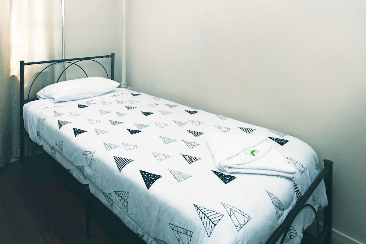 A single bed with a geometric-patterned bedspread in a room with a plain wall and a window with curtains.