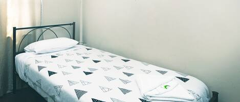 Una cama individual con una colcha de estampado geométrico en una habitación con pared lisa y una ventana con cortinas.
