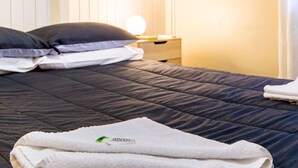 Ein ordentlich bezogenes Bett mit einer dunkelgrauen Decke, zwei Kissen und einem zusammengelegten weißen Handtuch daneben.