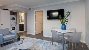 A modern living room with a flat-screen TV, a dining table with chairs, a grey sofa, a small coffee table, and a washing machine in the background. - 2BR The TMC Flats 04! Hermann Park Zoo! (Houston)