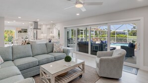 Una sala de estar moderna con sofá, sillón y mesa de centro, una cocina al fondo y un patio con vistas a una piscina.